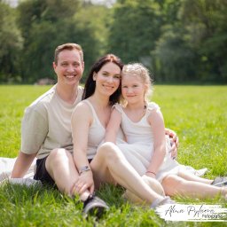 Alina Pylajewa - Uśmiechnięta rodzina (ojciec, matka i córka) siedząca na kocu na trawie w parku, w słoneczny dzień. Dziewczynka ma zaplecione warkocze. Na pierwszym planie widoczne buty sportowe.