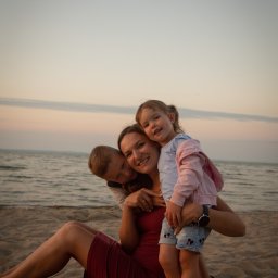 Alina Pylajewa - Matka siedzi na piaszczystej plaży, obejmując syna i córkę; w tle widać morze i niebo o zachodzie słońca, a dziewczynka ma na sobie dżinsowe szorty z motywem Myszki Miki.