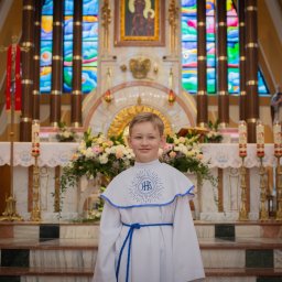 Alina Pylajewa - Chłopiec w stroju komunijnym na tle ołtarza z kwiatami i świecami, witraże w tle.