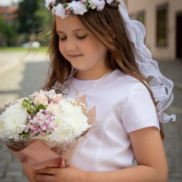 Alina Pylajewa - Dziewczynka w białej sukience trzyma bukiet kwiatów, z wiankiem na głowie i delikatnym naszyjnikiem, spogląda w dół na kwiaty na tle brukowanej drogi i budynku.