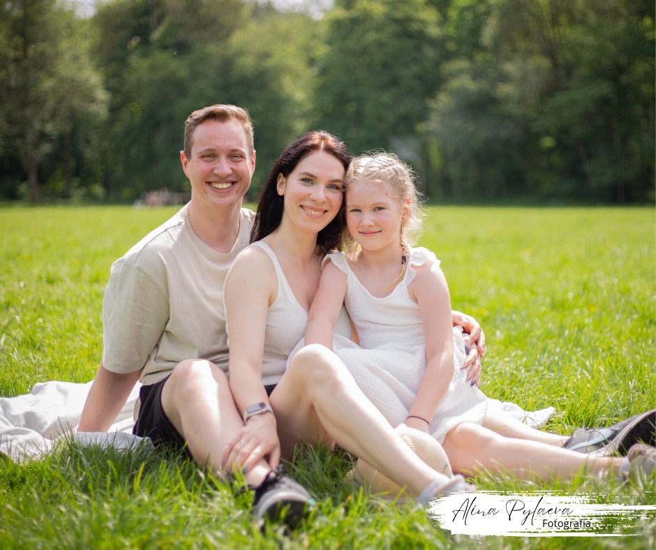 Uśmiechnięta rodzina (ojciec, matka i córka) siedząca na kocu na trawie w parku, w słoneczny dzień. Dziewczynka ma zaplecione warkocze. Na pierwszym planie widoczne buty sportowe.