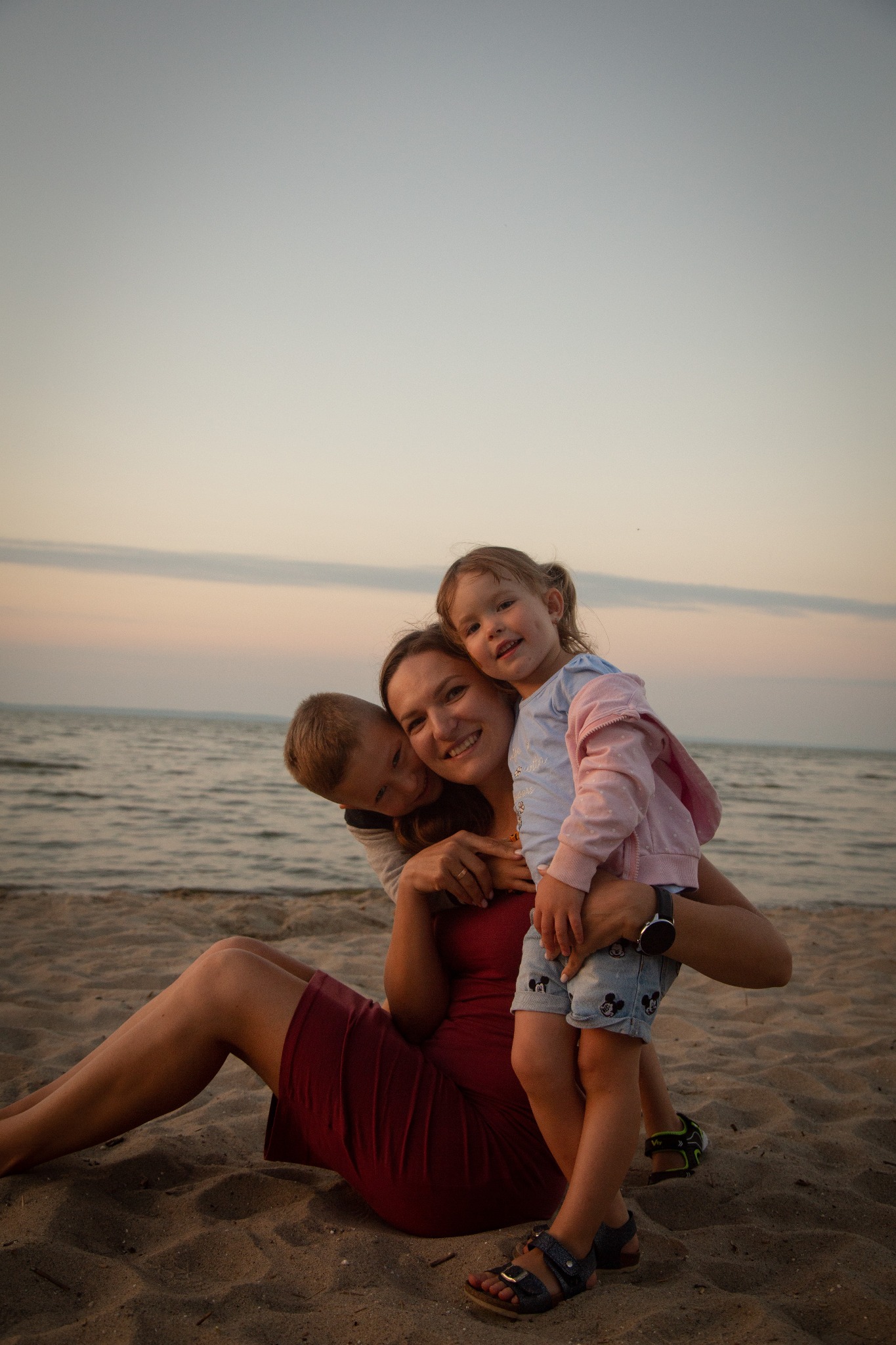 Matka siedzi na piaszczystej plaży, obejmując syna i córkę; w tle widać morze i niebo o zachodzie słońca, a dziewczynka ma na sobie dżinsowe szorty z motywem Myszki Miki.