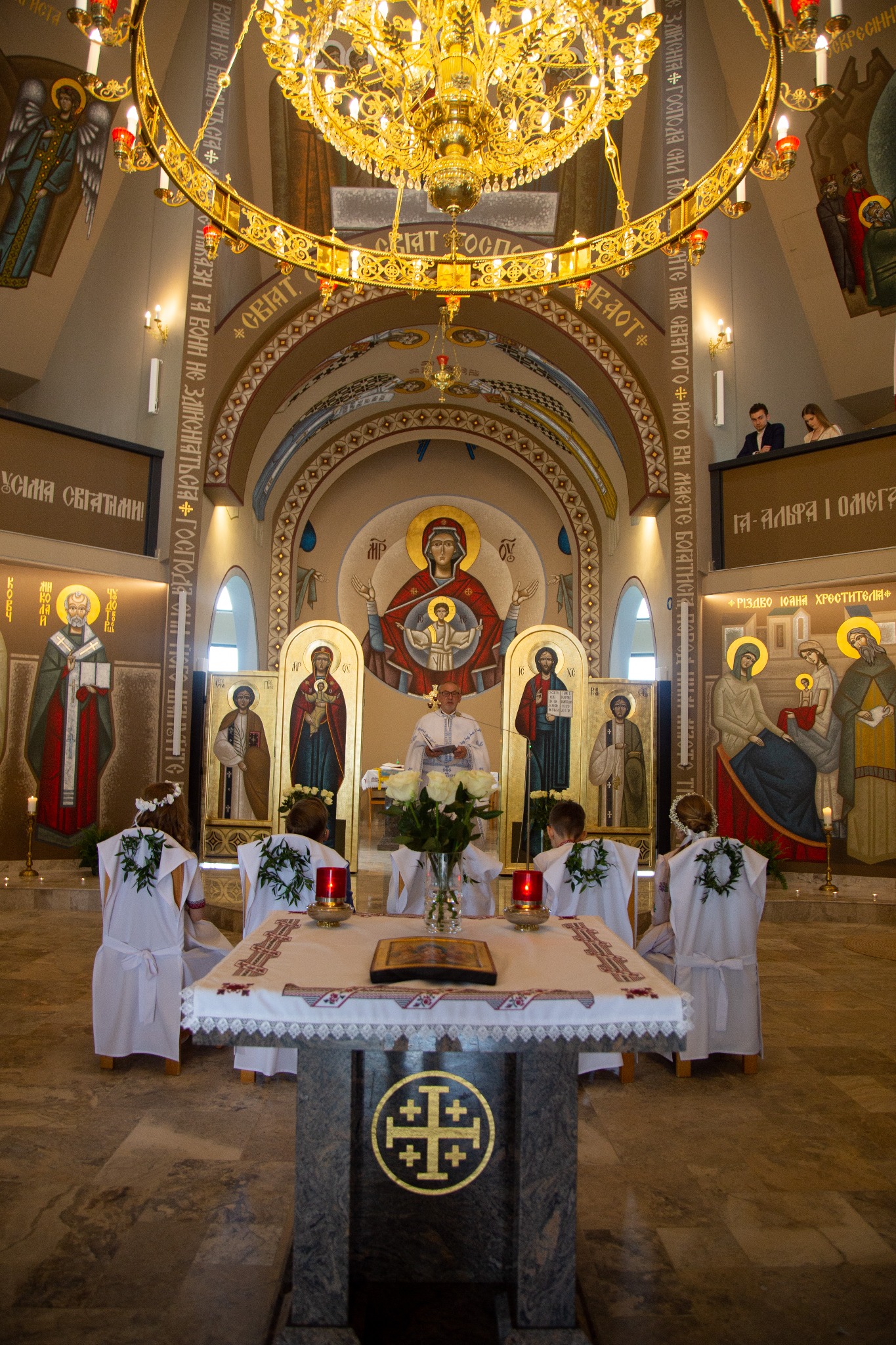 Wnętrze cerkwi podczas uroczystości religijnej. Dzieci w białych szatach z wiankami na głowach stoją przed ołtarzem. Ksiądz w białej szacie prowadzi ceremonię. W tle ikony i złoty żyrandol.