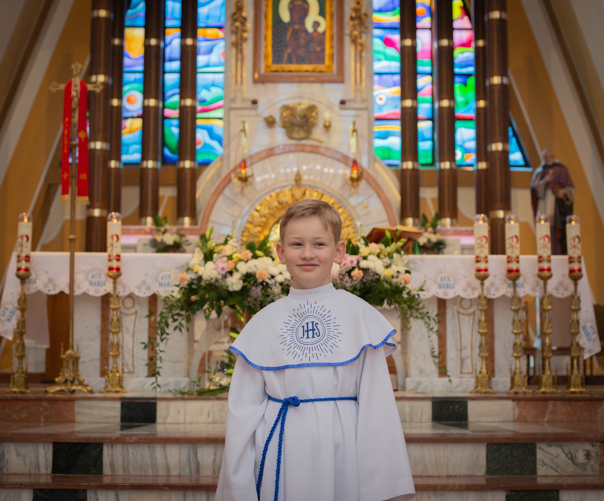 Chłopiec w stroju komunijnym na tle ołtarza z kwiatami i świecami, witraże w tle.
