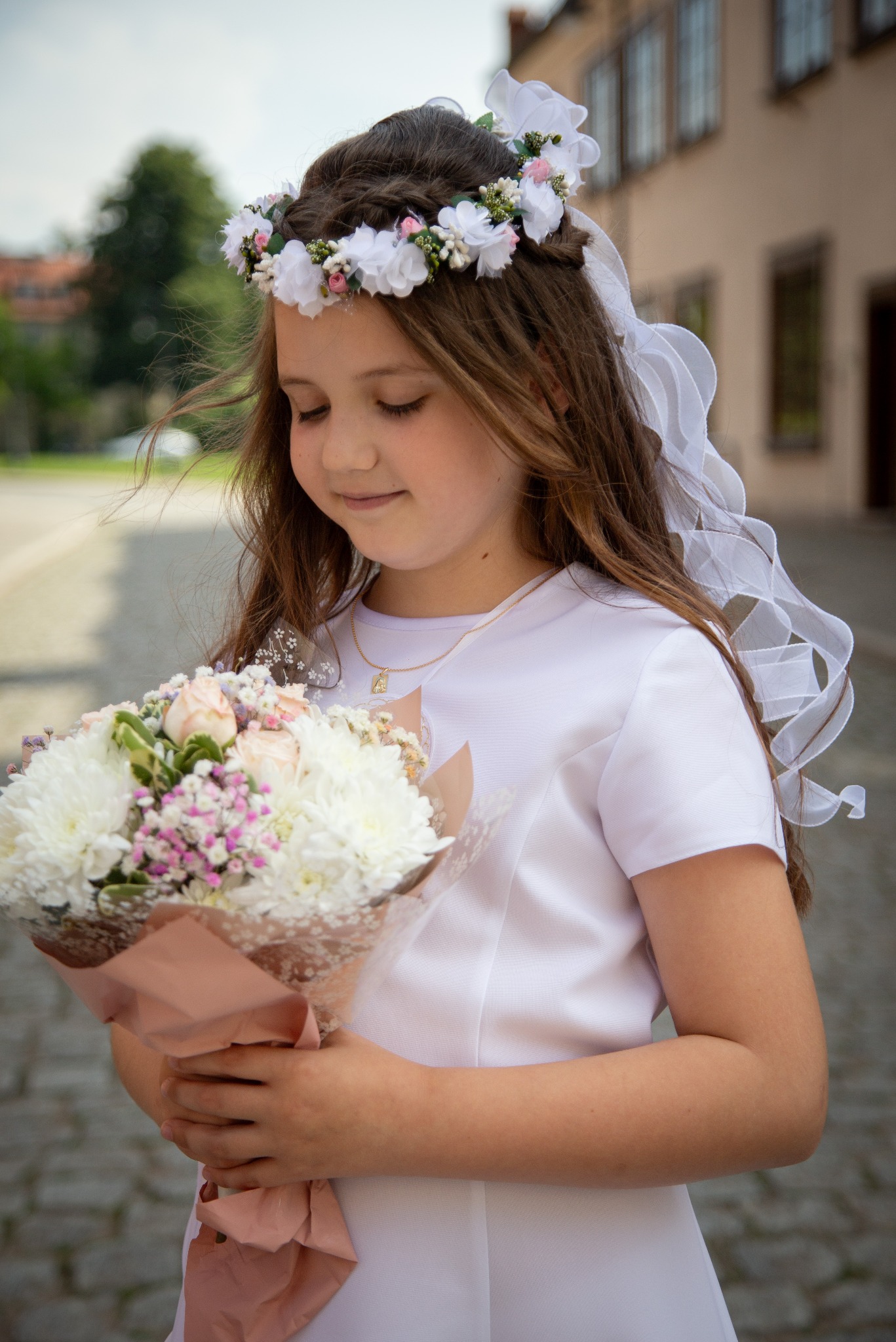 Dziewczynka w białej sukience trzyma bukiet kwiatów, z wiankiem na głowie i delikatnym naszyjnikiem, spogląda w dół na kwiaty na tle brukowanej drogi i budynku.