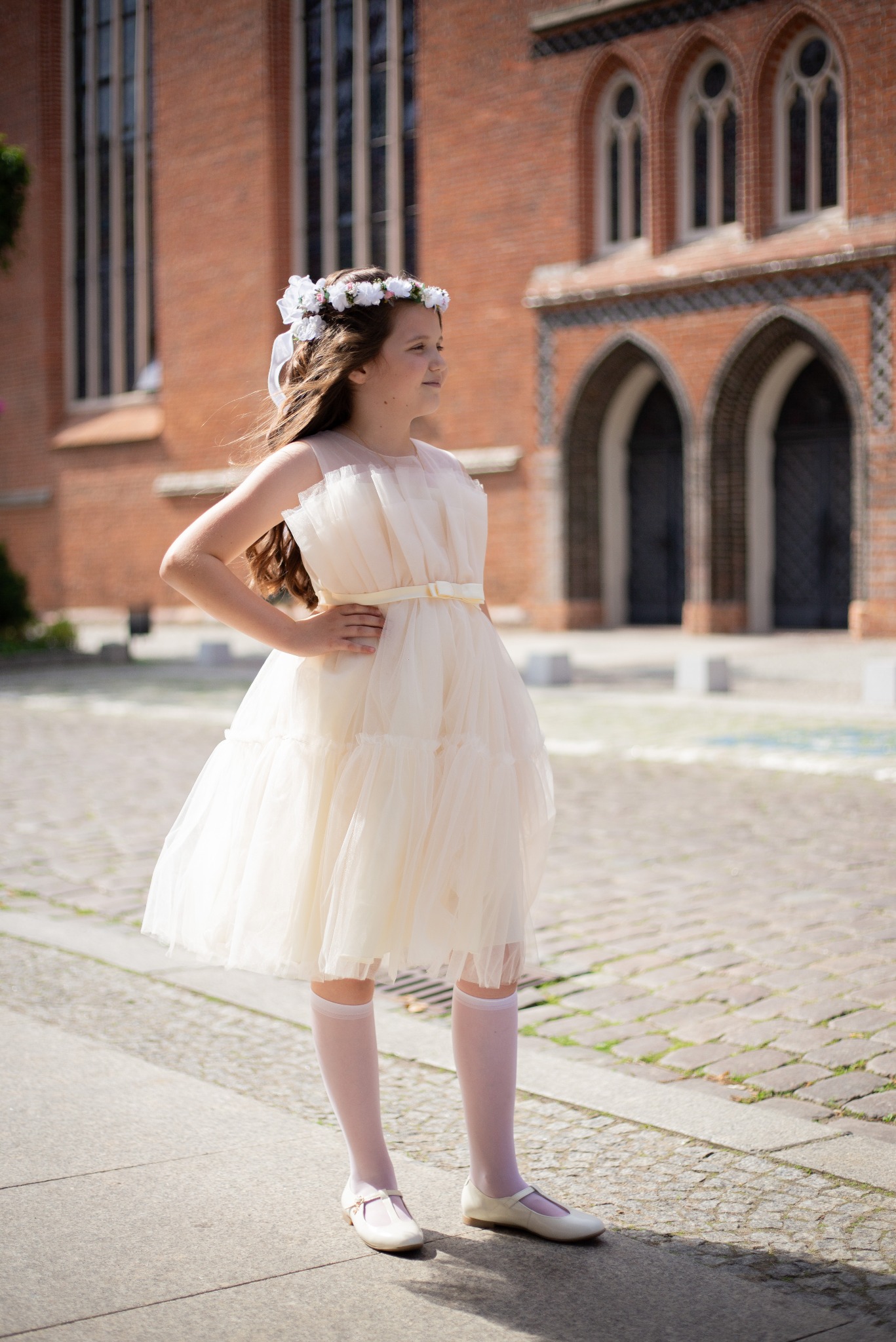 Dziewczynka w kremowej sukience z tiulu i wiankiem z białych kwiatów na głowie, pozuje na tle ceglanego budynku z gotyckimi oknami.