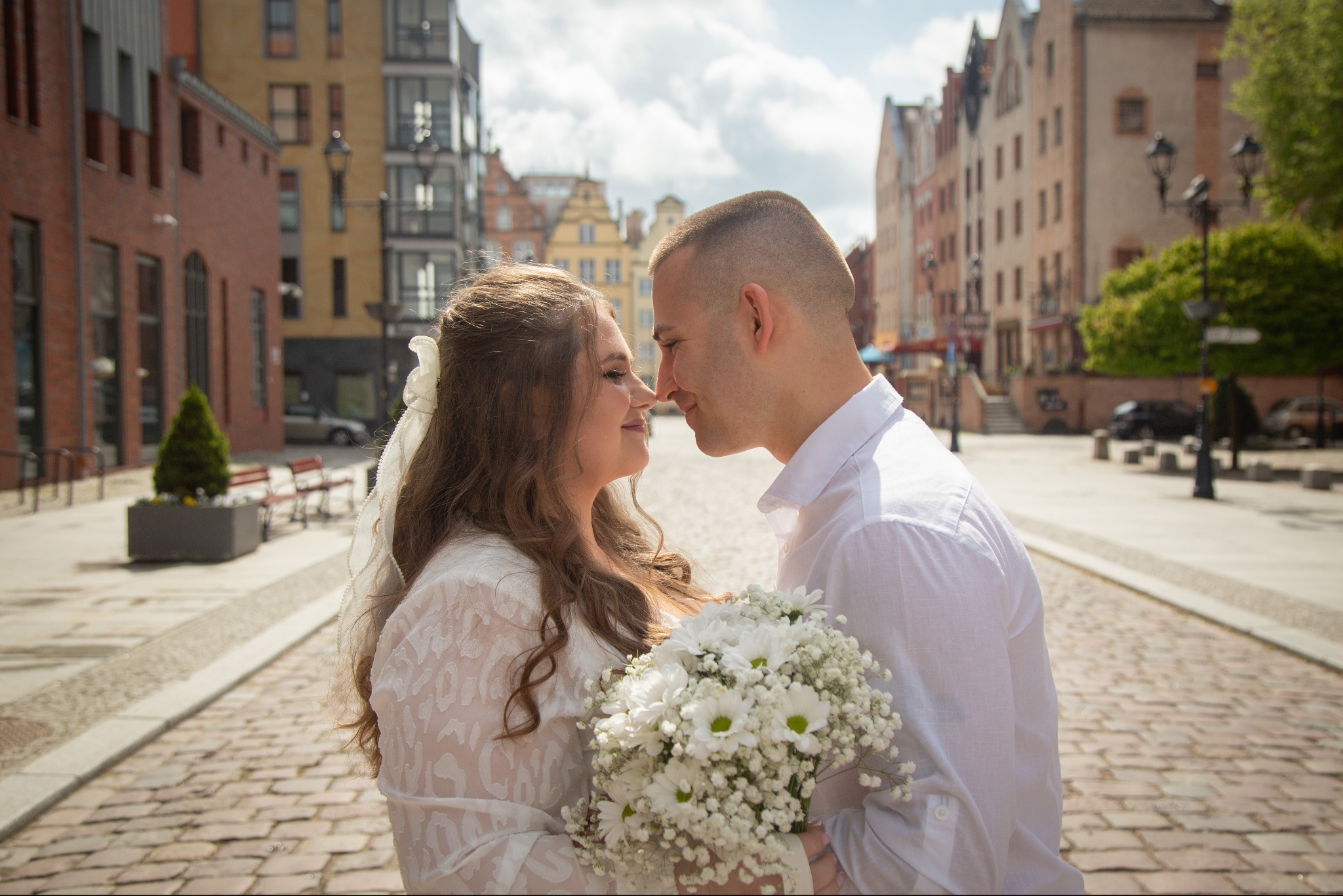 Para młoda, delikatnie dotykająca się nosami na brukowanej uliczce w historycznym mieście, panna młoda trzyma bukiet białych kwiatów, w tle budynki z czerwonej cegły i kamienice.