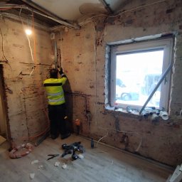 U.R.B GK SYSTEM - Elektryk w kamizelce odblaskowej montuje rurę na ścianie z odsłoniętą cegłą w trakcie prac instalacyjnych; widoczne przewody, narzędzia i okno z widokiem na zewnątrz.