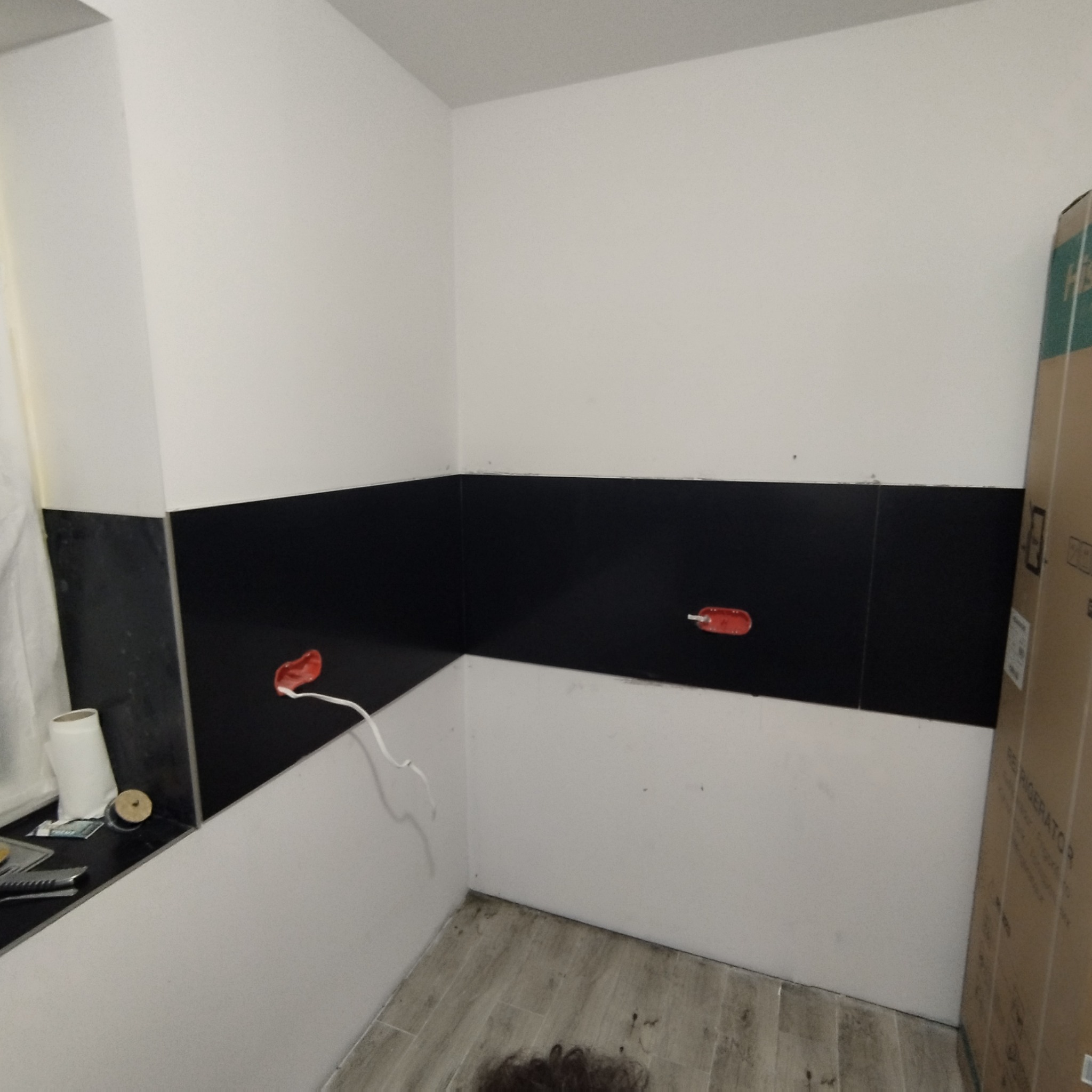 Łazienka w trakcie prac wykończeniowych: dolna część ścian wyłożona czarnymi płytkami, górna część biała, widoczne puszki elektryczne i przewody, podłoga z płytek imitujących drewno, kartonowe...