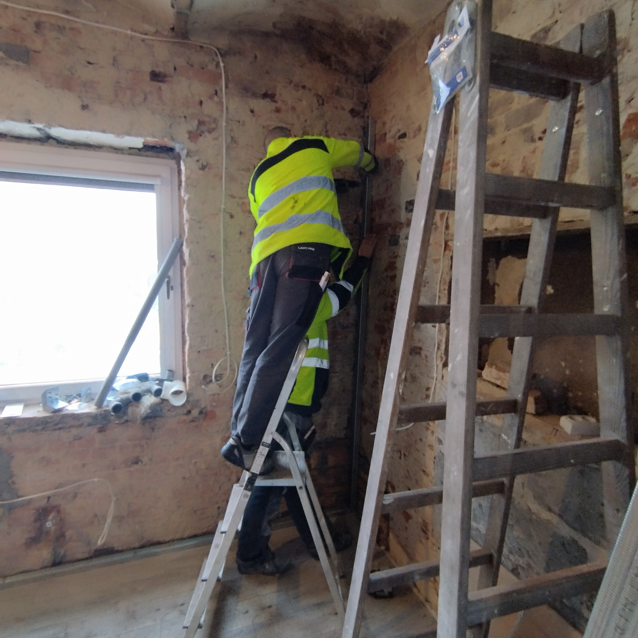 Dwóch pracowników w odblaskowych kamizelkach montuje profile na ceglanej ścianie w trakcie remontu, widoczne drabiny, okno z rurami i narzędziami na parapecie.