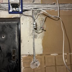 Jacek Klimek - Szczegółowe ujęcie licznika elektrycznego z podłączonymi przewodami na ścianie w trakcie prac instalacyjnych. Widoczne kable i elementy montażowe.