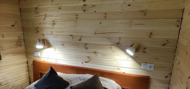 Drewniana sypialnia z dwoma kinkietami o ciepłym świetle, zamontowanymi nad wezgłowiem łóżka, obok gniazdek elektrycznych.