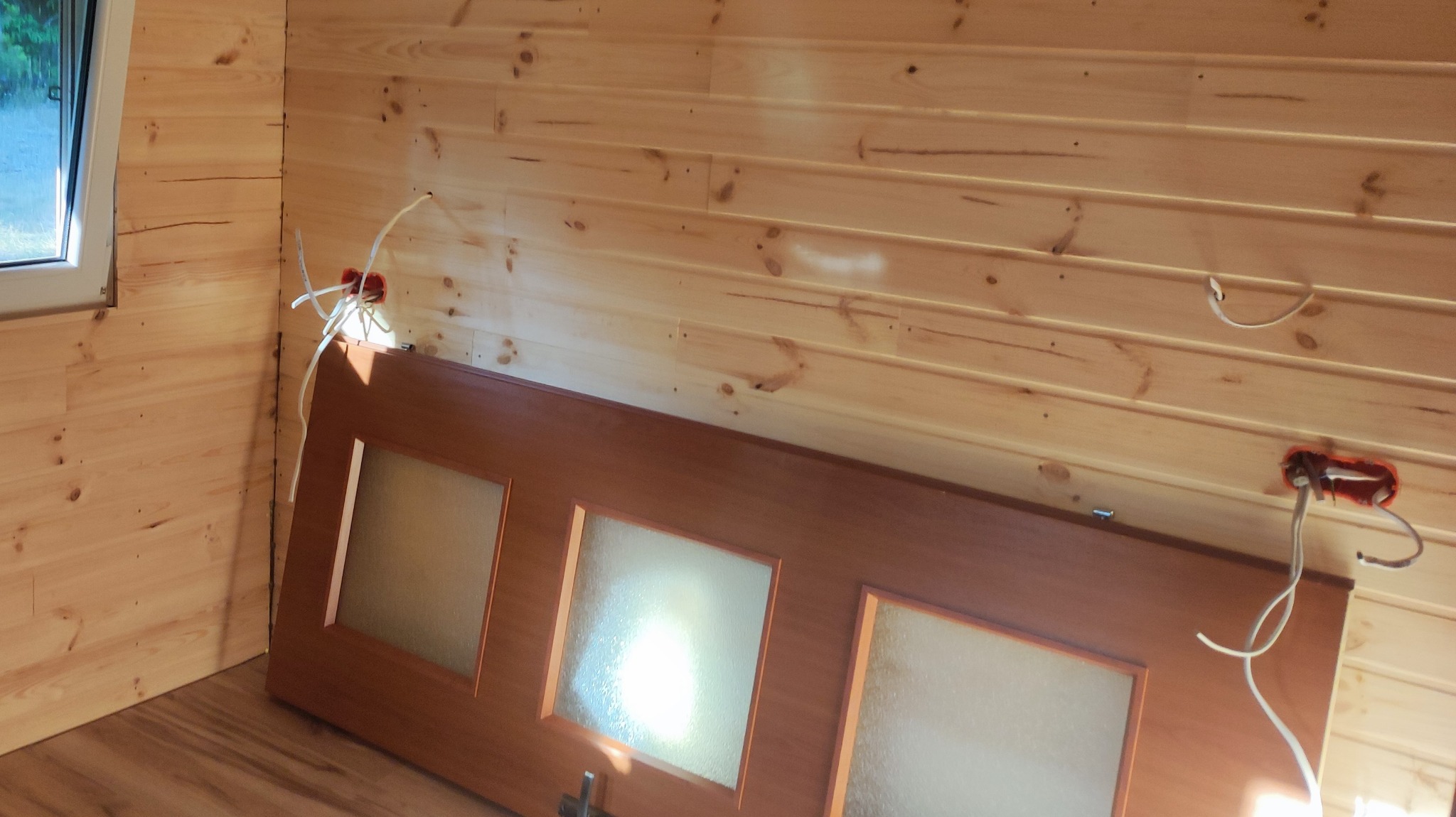 Wnętrze wykończone boazerią, z wystającymi przewodami elektrycznymi ze ściany i sufity, oraz opartymi o ścianę brązowymi drzwiami z trzema przeszkleniami.