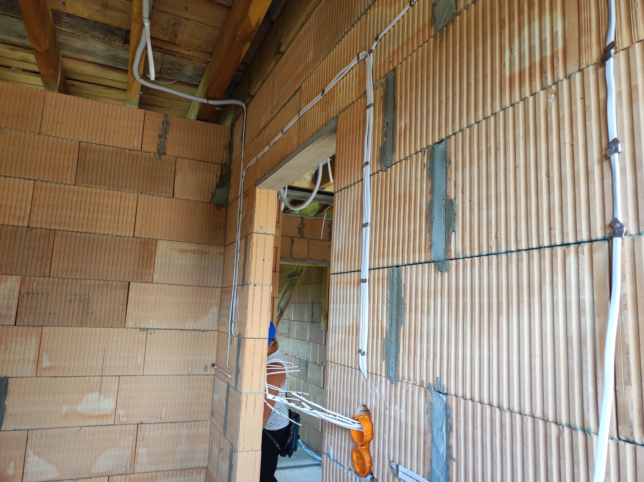 Instalacja elektryczna w surowym budynku: białe kable prowadzone po ścianie z cegieł ceramicznych, mocowane metalowymi uchwytami, widoczne puszki instalacyjne i fragment osoby w tle.