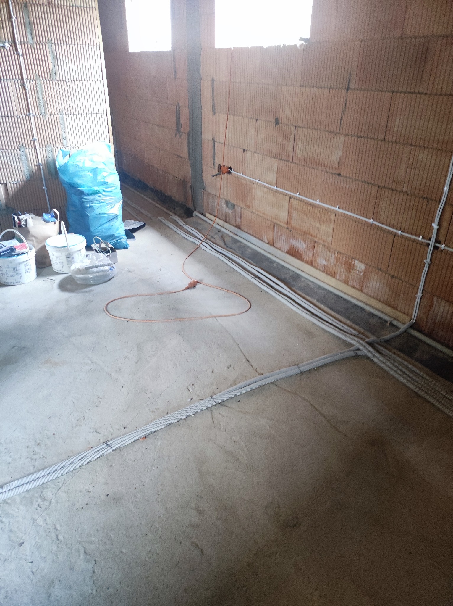 Wnętrze w trakcie prac wykończeniowych, widoczne kable elektryczne prowadzone po ścianach z cegły oraz rury instalacyjne na podłodze, obok stoją wiadra i niebieski worek na śmieci.