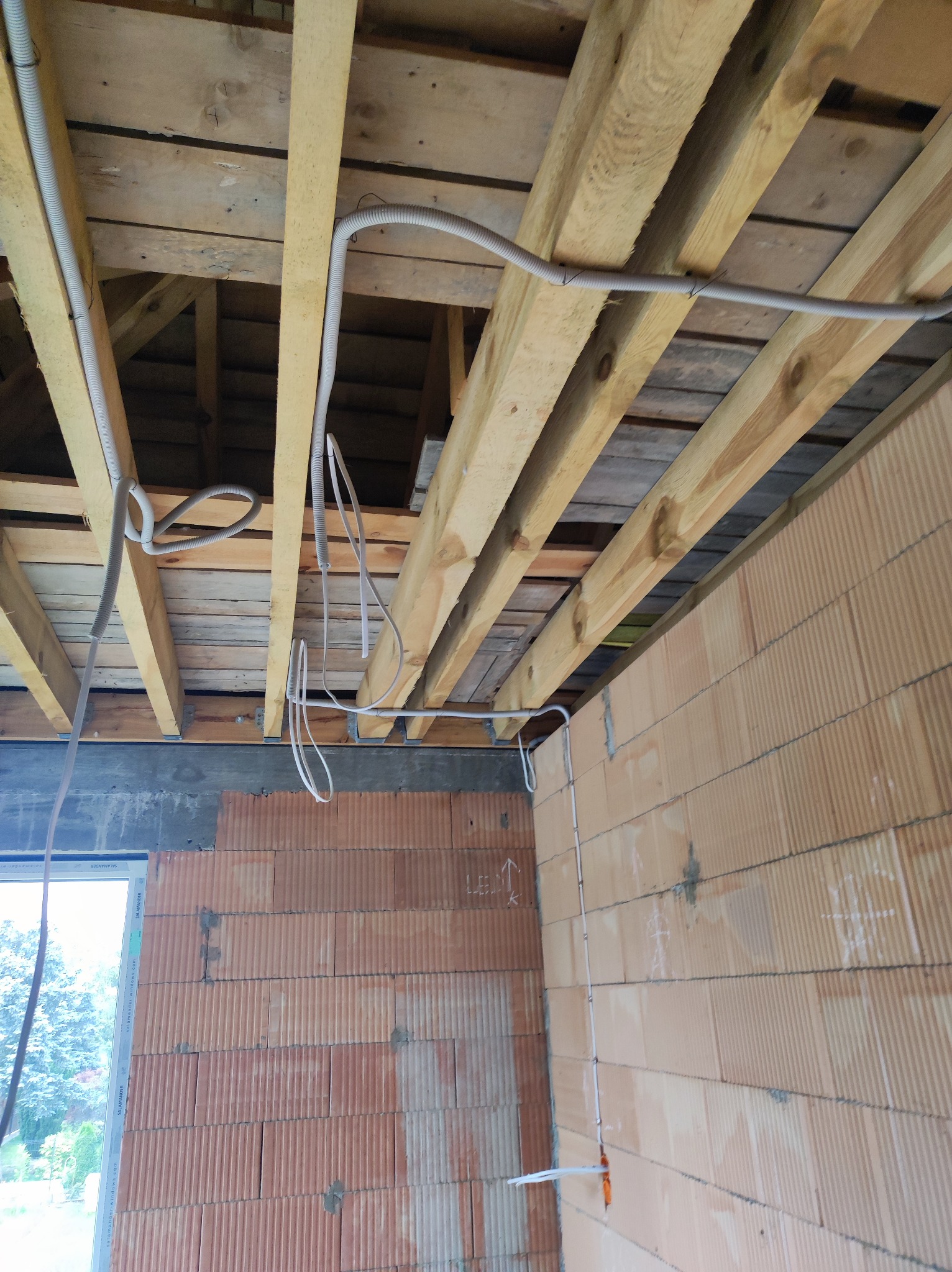 Wnętrze w trakcie budowy: widok na sufit z drewnianą konstrukcją i częściowo ułożonymi kablami w peszlach, ściany z cegły ceramicznej, okno z widokiem na zewnątrz.