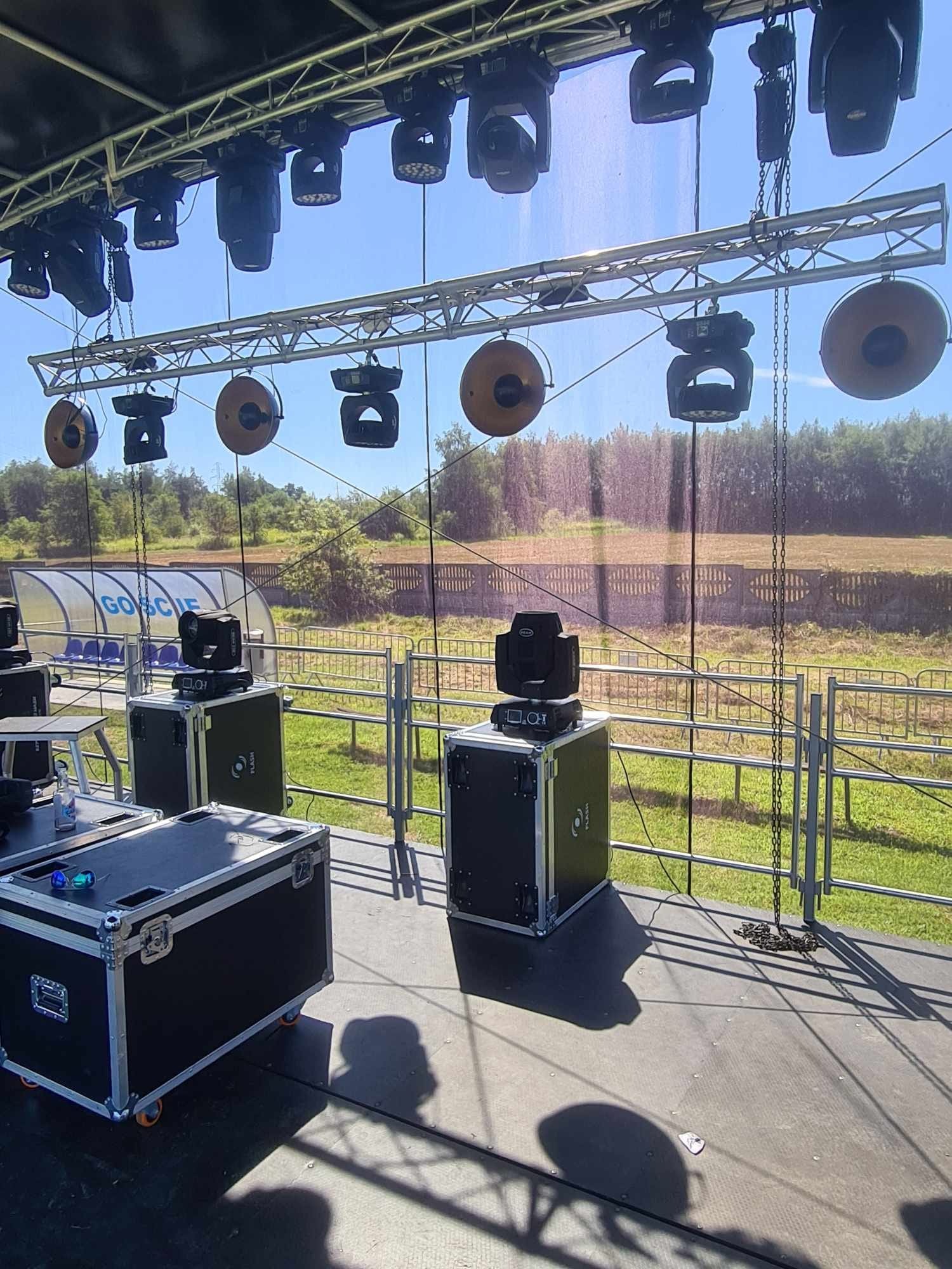 Scena plenerowa z zamontowanym oświetleniem na metalowej konstrukcji kratownicowej, reflektory punktowe i okrągłe, w tle widoczne drzewa i trybuna dla kibiców, sprzęt oświetleniowy w czarnych...