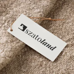 Biała metka z czarnym logo przedstawiającym sukienkę i napisem 'szatoland' leży na beżowej, teksturowanej tkaninie o pętelkowej strukturze.