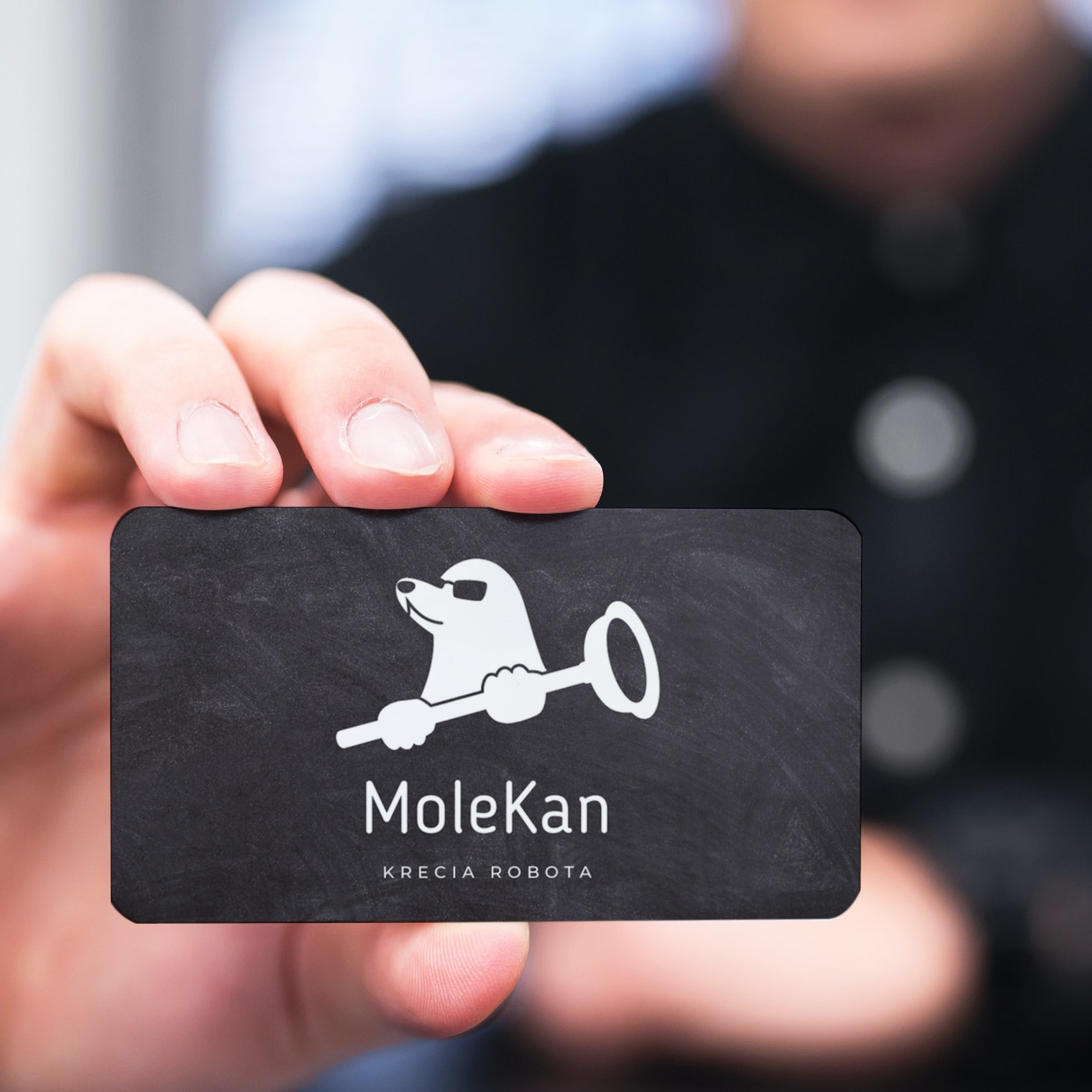 Osoba trzyma czarną wizytówkę z białym logo kreta w okularach słonecznych trzymającego łopatę i nazwą firmy MoleKan KRECIA ROBOTA.