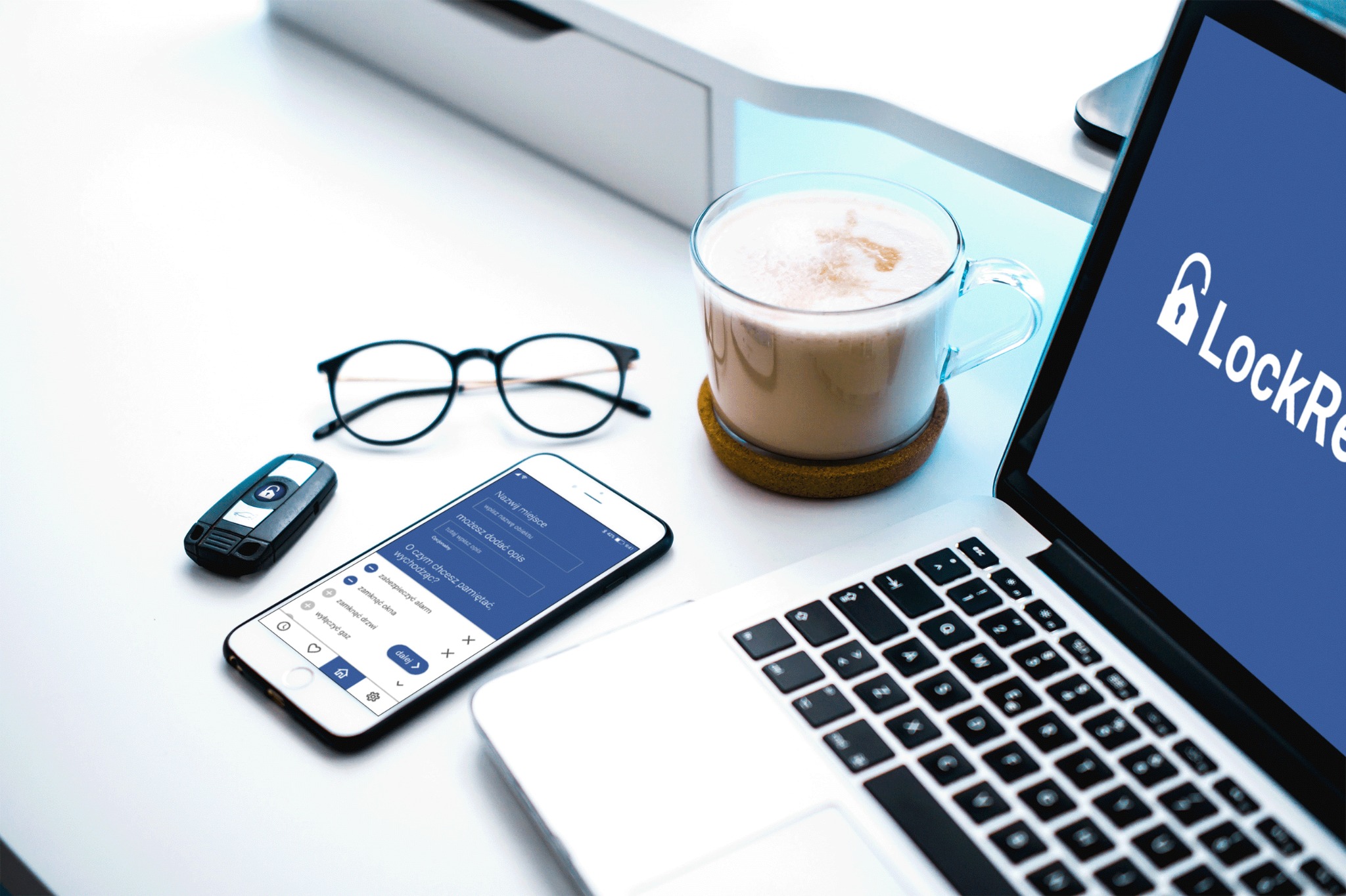 Laptop z logo LockRe, smartfon z interfejsem aplikacji do zarządzania bezpieczeństwem domu, okulary, brelok samochodowy z symbolem kłódki i kawa na biurku.