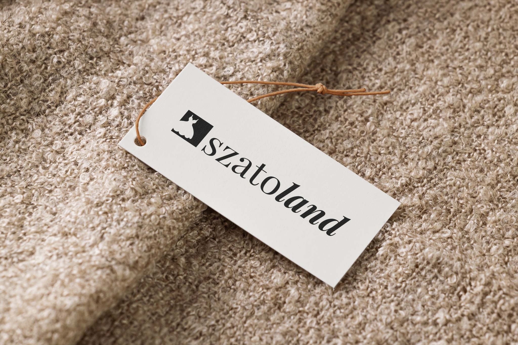 Biała metka z czarnym logo przedstawiającym sukienkę i napisem 'szatoland' leży na beżowej, teksturowanej tkaninie o pętelkowej strukturze.