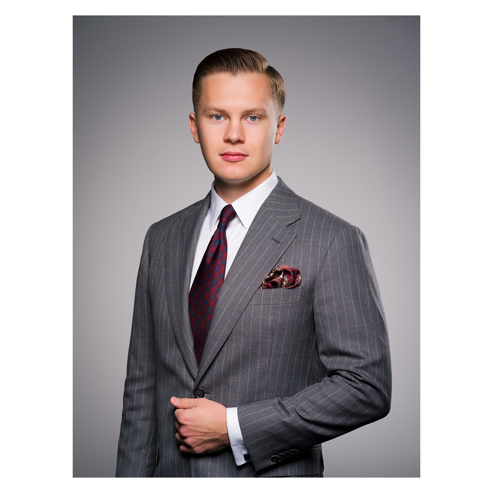 Portret mężczyzny w szarym garniturze w prążki, z bordowym krawatem w geometryczny wzór i ozdobną poszetką w kieszonce, na jednolitym szarym tle.