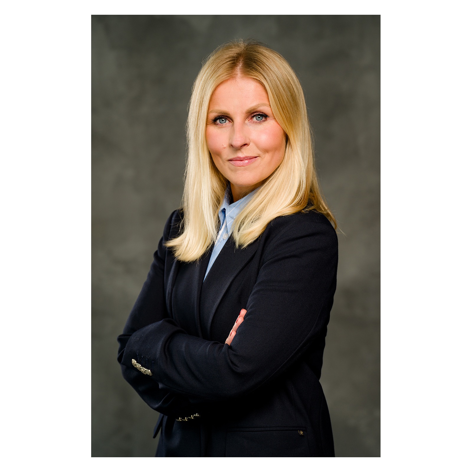 Portret kobiety z blond włosami i niebieskimi oczami, ubrana w granatową marynarkę i błękitną koszulę, z założonymi rękami na ciemnoszarym tle.