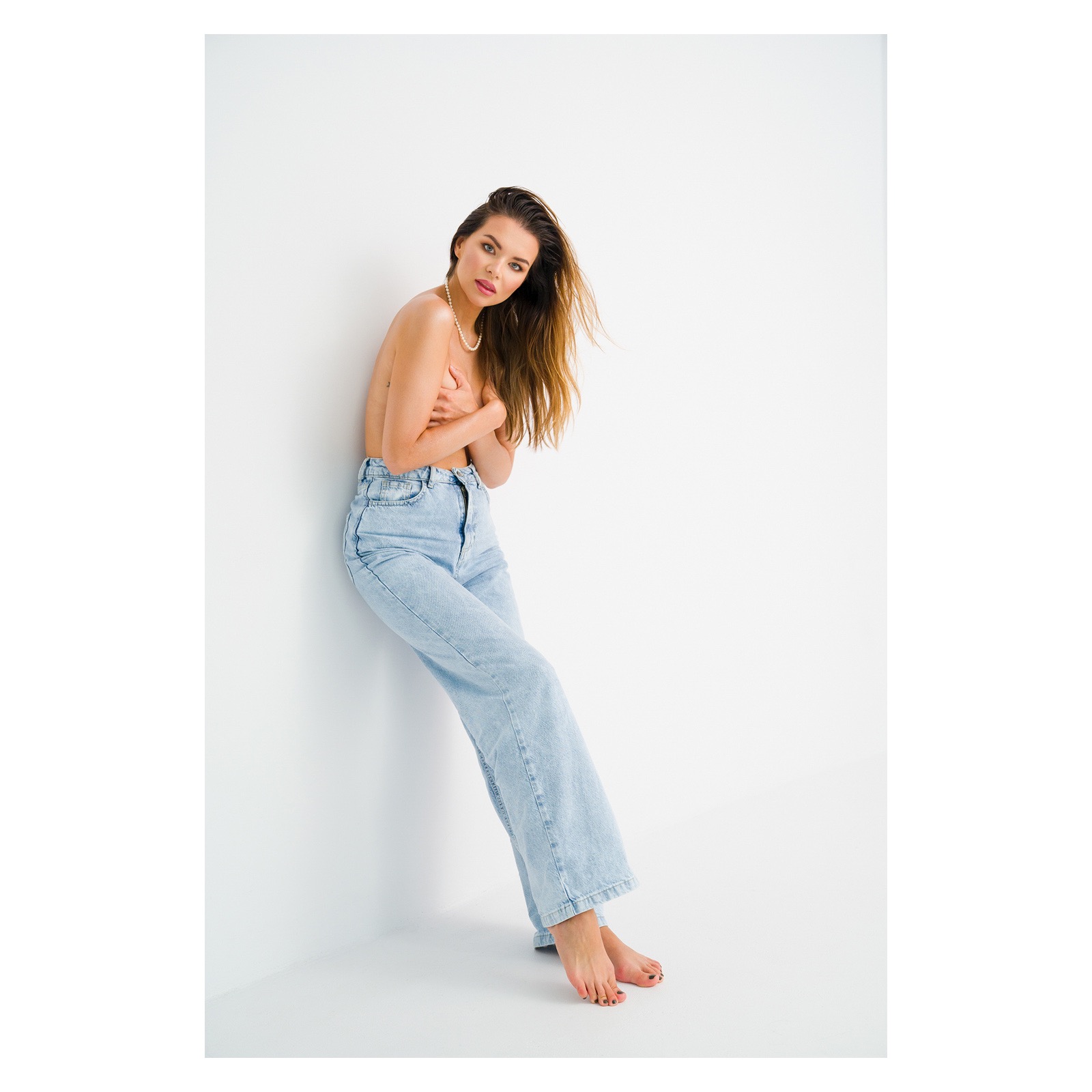 Portret kobiety opierającej się o białą ścianę, z mokrymi włosami, w dżinsach i z naszyjnikiem z pereł, zasłaniającej tors rękami.