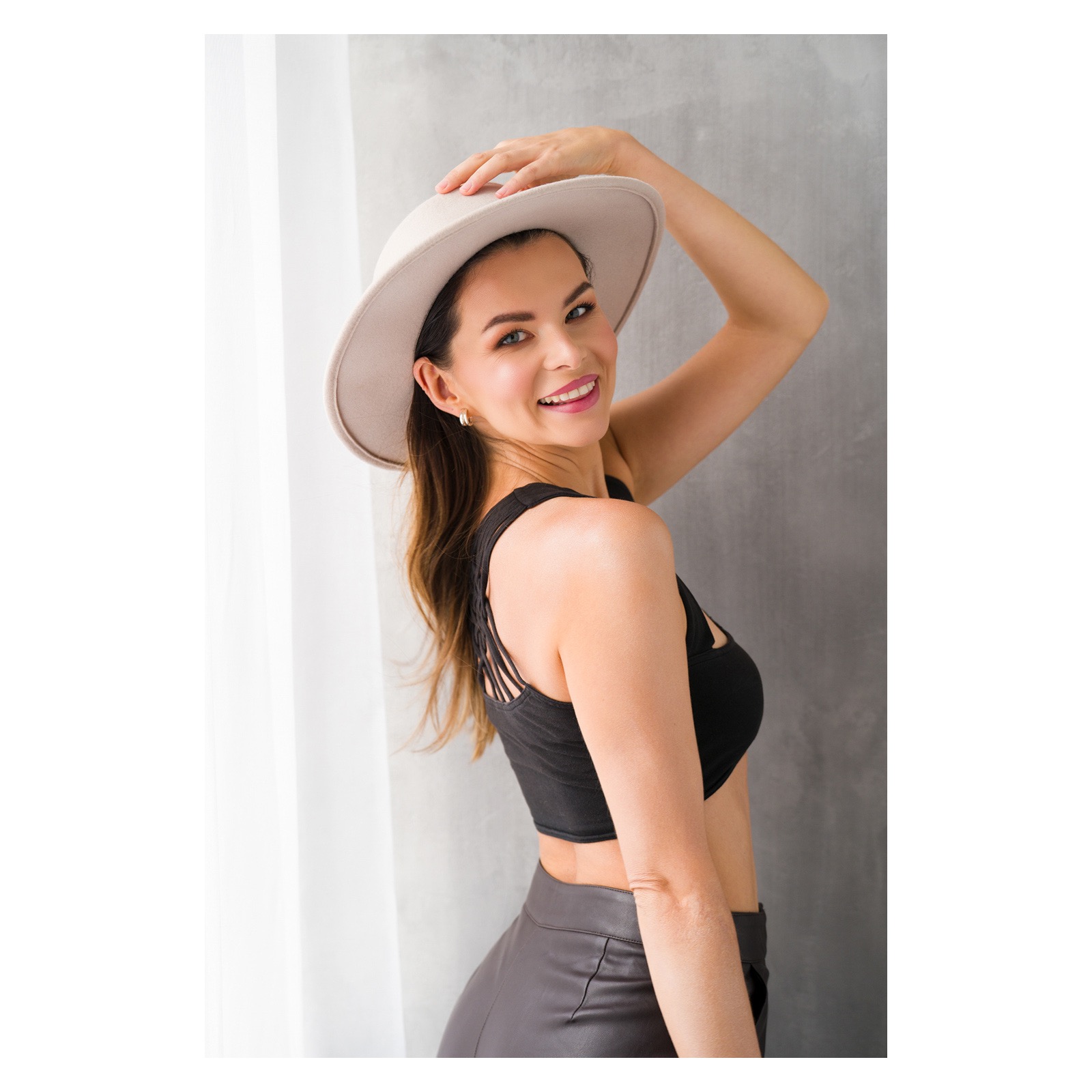 Uśmiechnięta kobieta w kapeluszu, czarnym topie i skórzanych spodniach pozuje na tle szarej ściany i białej zasłony.