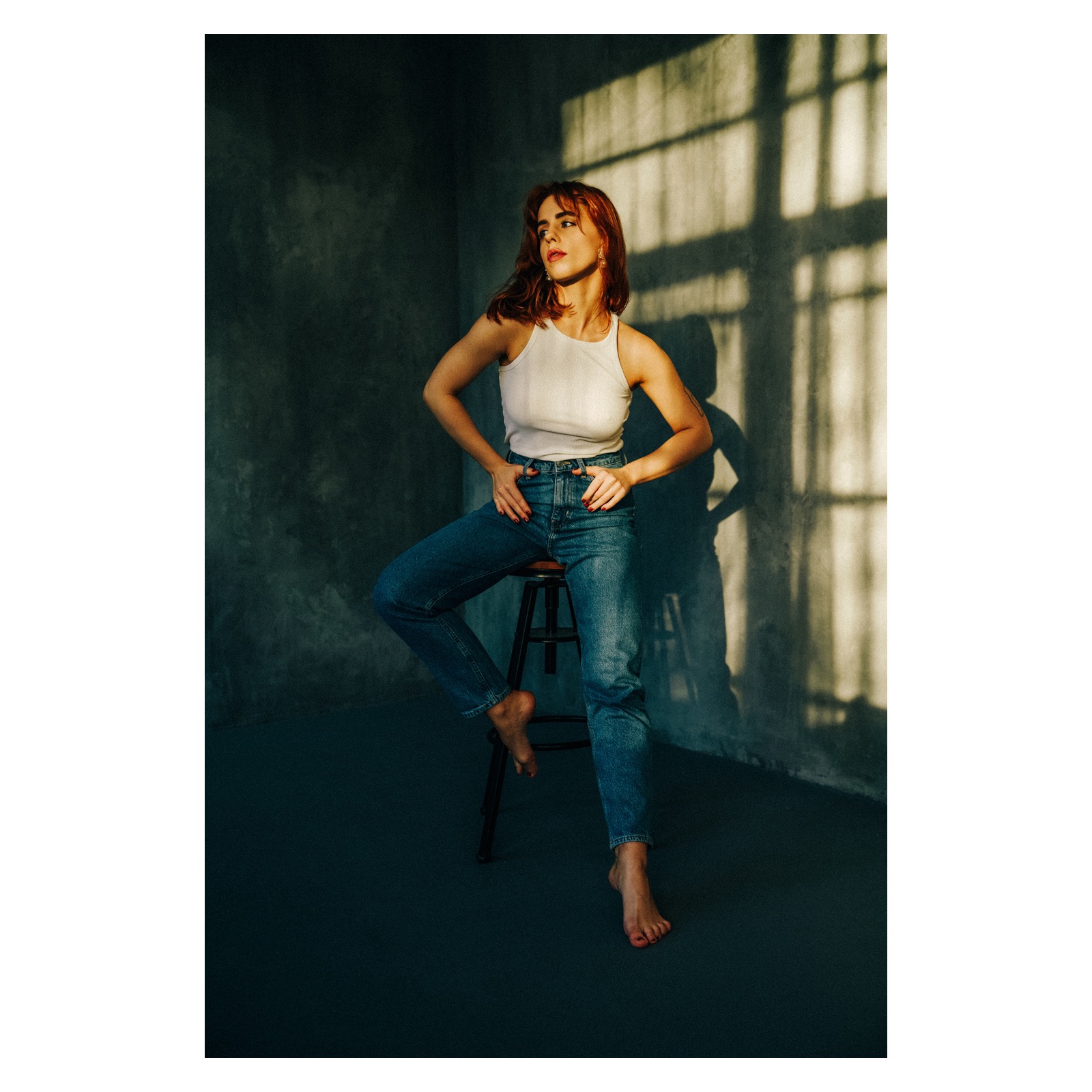 Kobieta z rudymi włosami siedzi na stołku w dżinsach i białym topie na tle szarej ściany z cieniem okna, z rękami opartymi na biodrach.