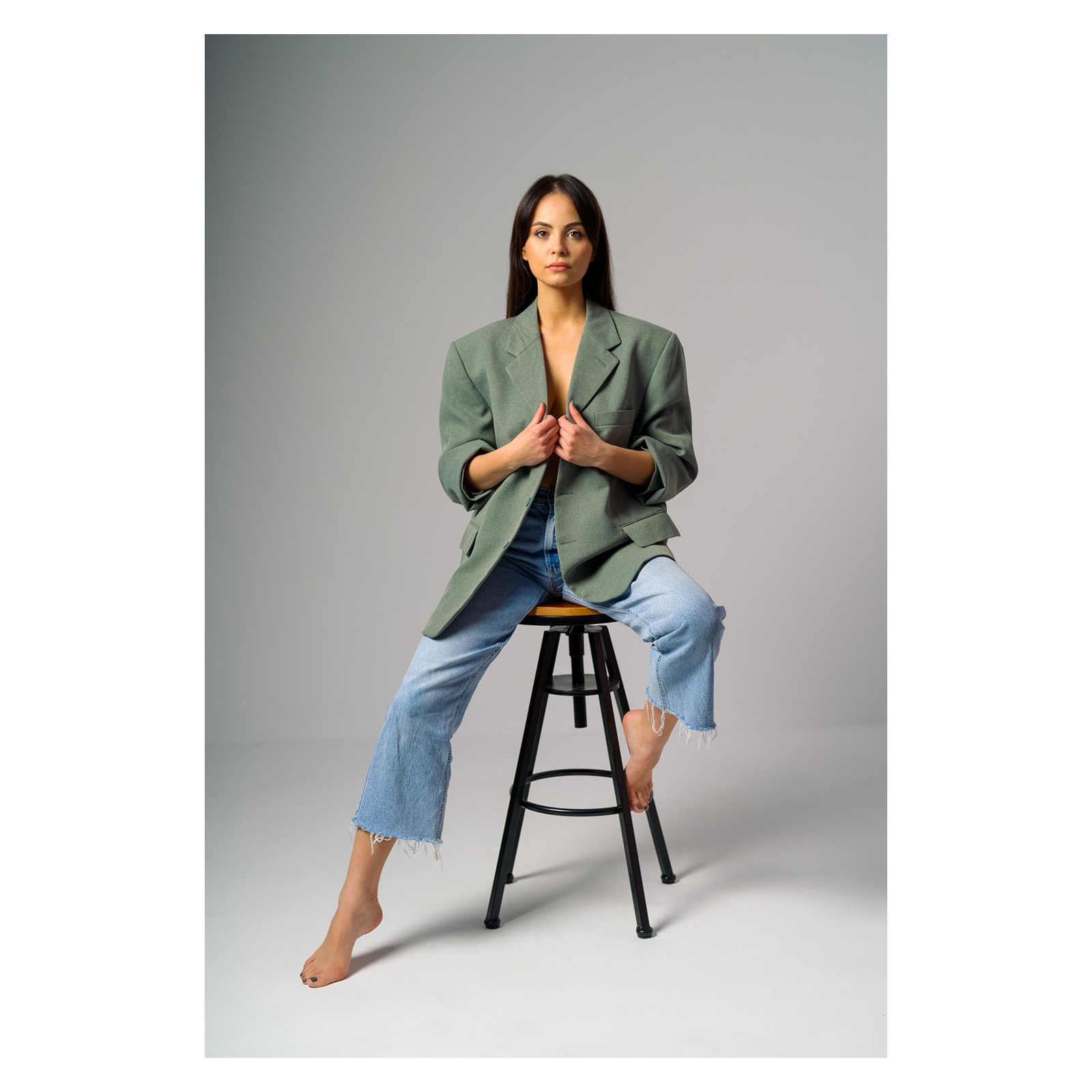 Kobieta siedząca na stołku, ubrana w dżinsy z postrzępionymi nogawkami i oversize'ową zieloną marynarkę, pozująca na szarym tle.