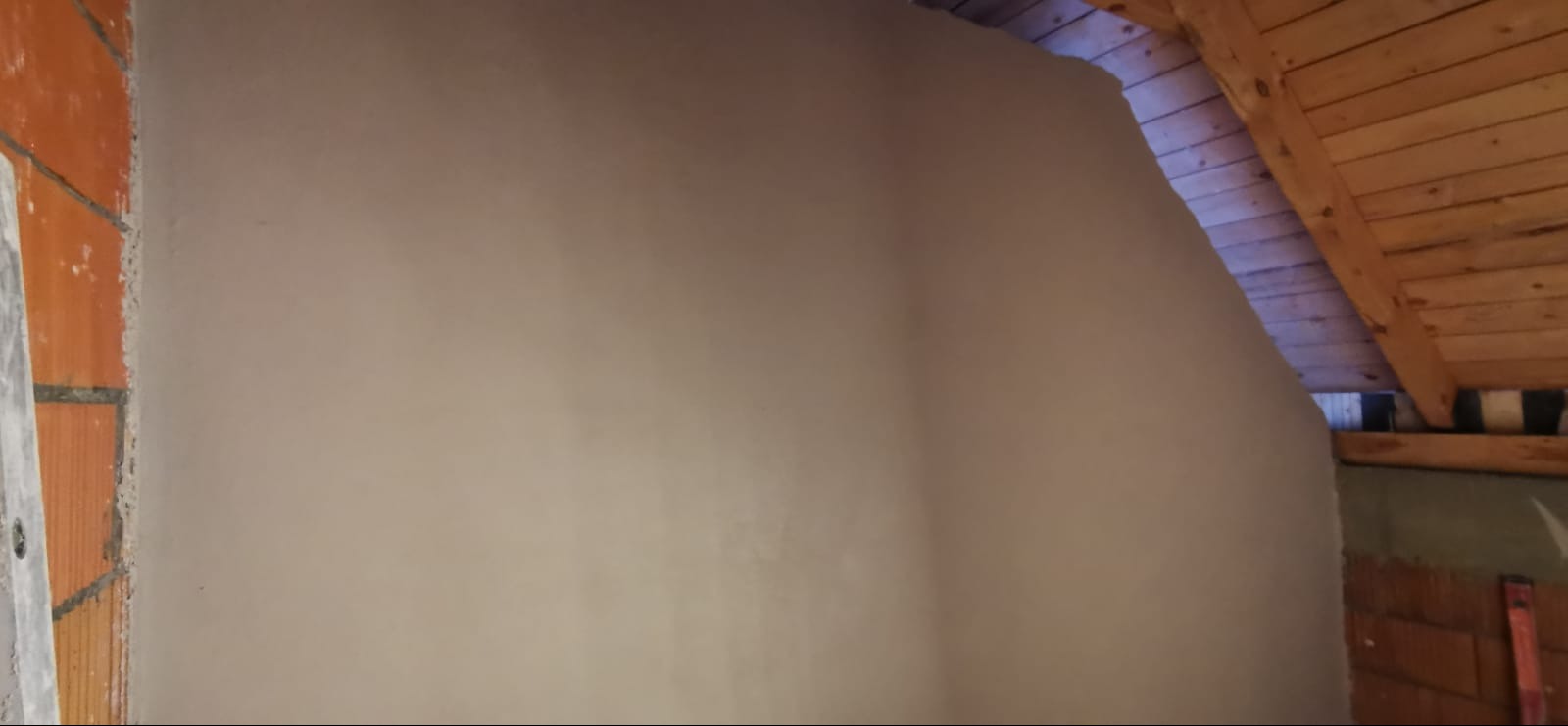 Świeżo nałożony tynk maszynowy na ścianie poddasza, widoczne drewniane elementy konstrukcji dachu i fragment muru z cegły.