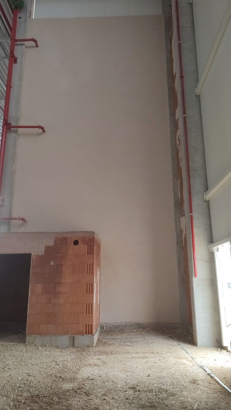 Wysoka ściana pokryta świeżym, jasnym tynkiem w surowym wnętrzu, widoczne pionowe rury instalacyjne w kolorze czerwonym, fragment ściany z cegły dziurawki i betonowe elementy konstrukcyjne.