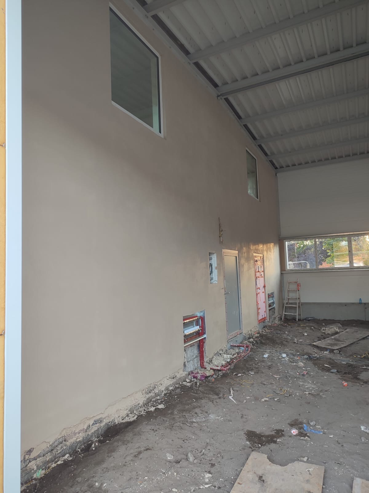 Wnętrze hali w trakcie prac wykończeniowych: ściana po nałożeniu beżowego tynku, widoczne okna, instalacje elektryczne w ścianie, fragment metalowej konstrukcji dachu i bałagan na podłodze.