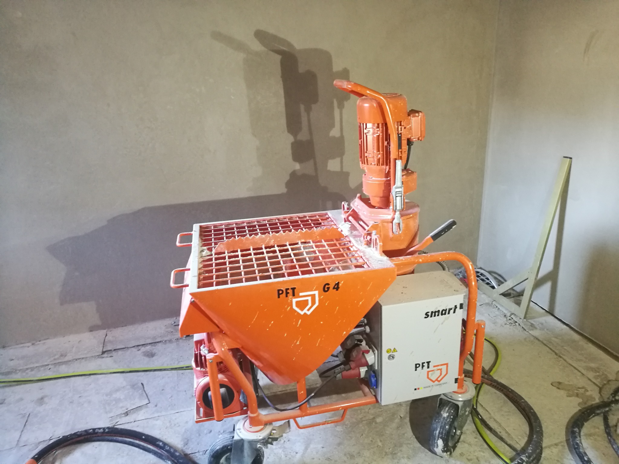 Pomarańczowa maszyna do tynkowania PFT G4 Smart stoi na podłodze w pokoju z częściowo otynkowanymi ścianami, widoczny cień maszyny na ścianie.