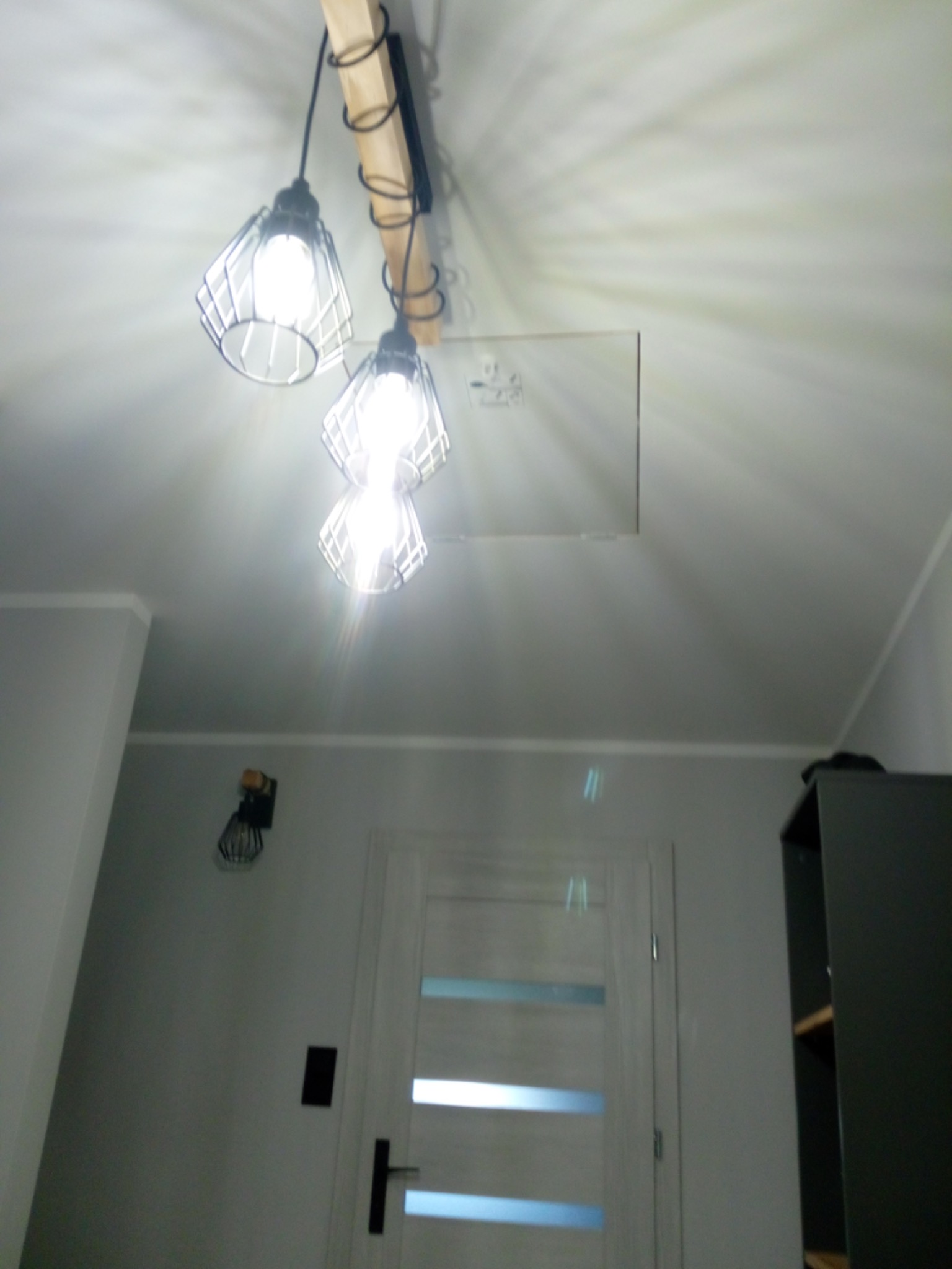 Nowoczesna lampa wisząca z trzema kloszami w kształcie klatek, zamontowana na drewnianej belce, oświetlająca białe wnętrze z drzwiami z przeszkleniami i szafką.