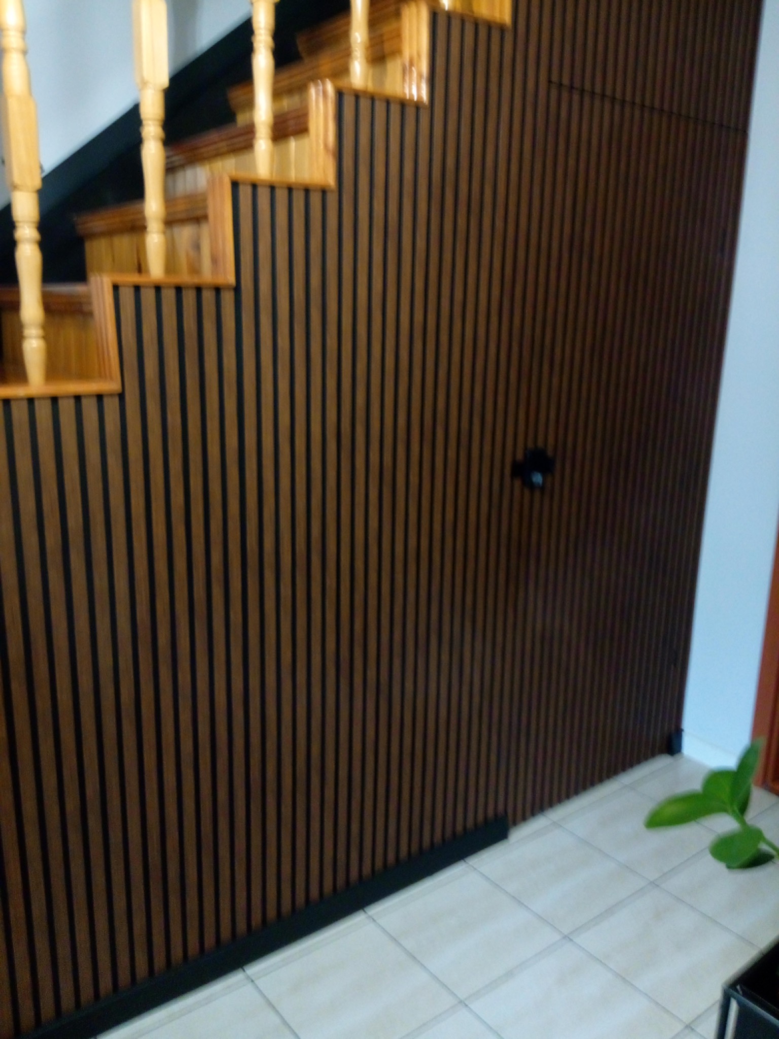 Drewniane panele ścienne z ciemnymi listwami tworzące pionowy wzór, dopasowane do drewnianych schodów z jasnymi balustradami, na podłodze jasne płytki.