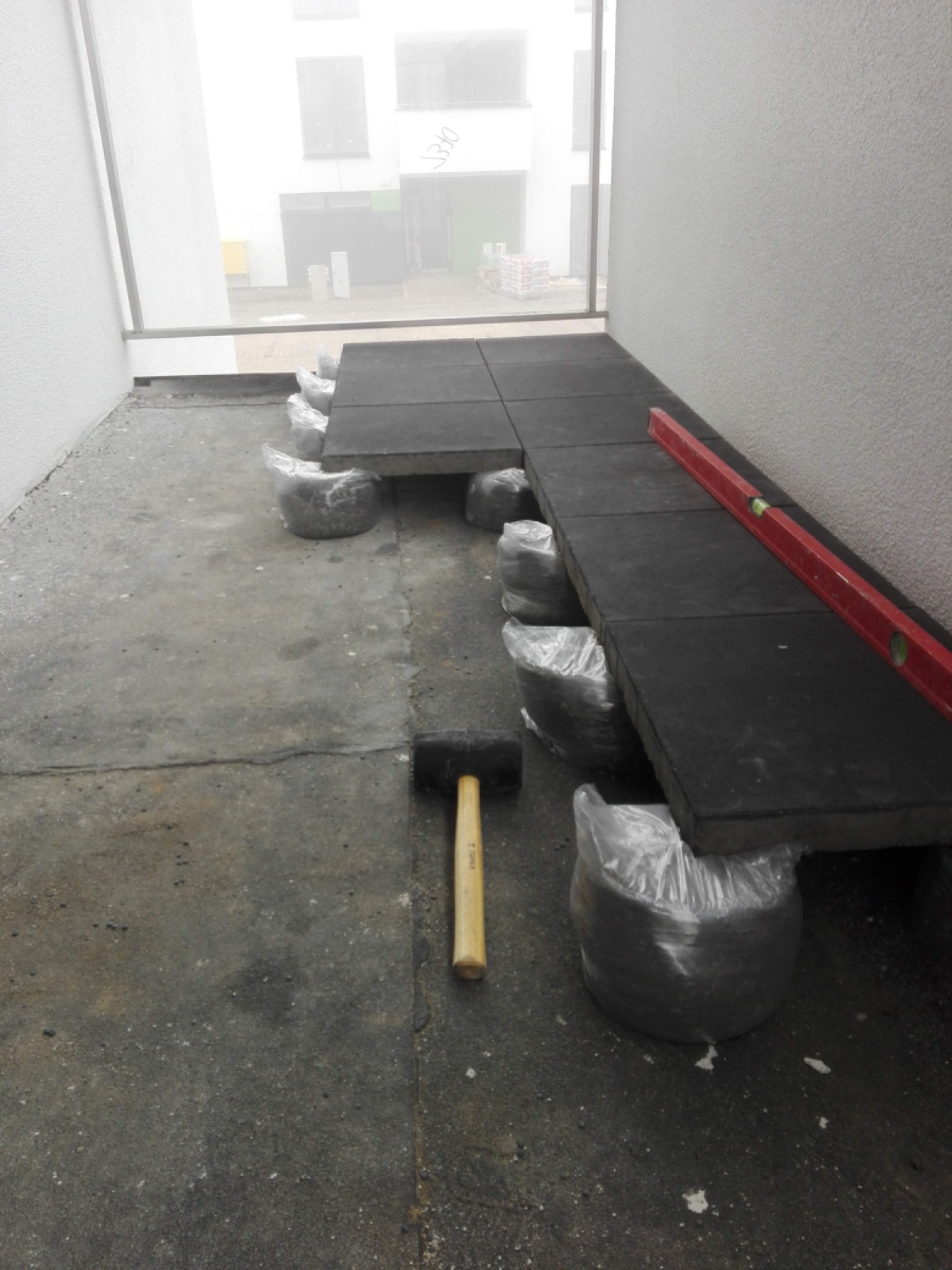 Montaż czarnych płyt podłogowych na regulowanych podporach z worków foliowych, poziomica i młotek gumowy na szarym podłożu, widok przez okno na budynek w tle.