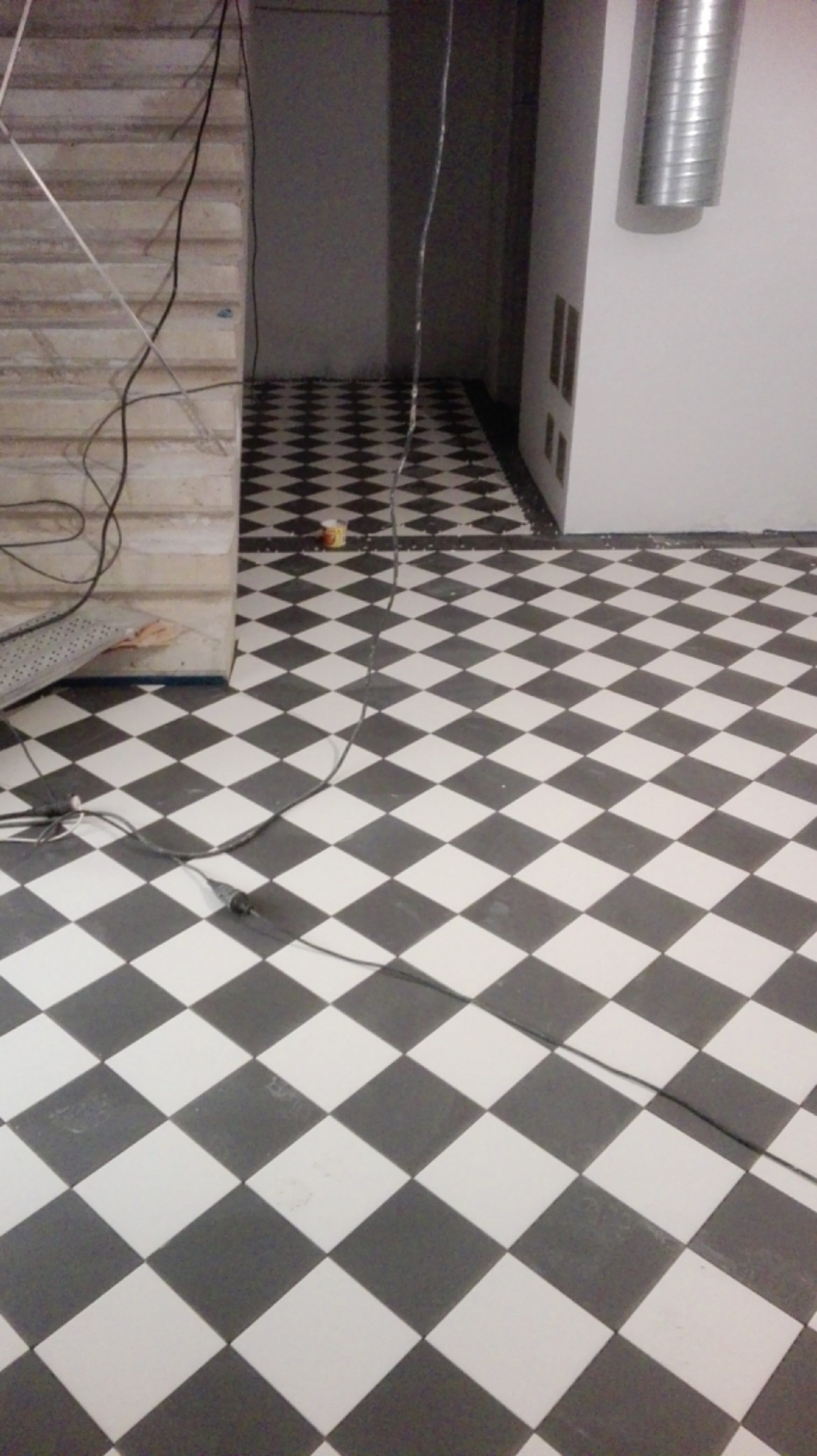 Ułożona podłoga z czarno-białych płytek w romby, widoczne kable i puszka na podłodze, fragment schodów w tle.
