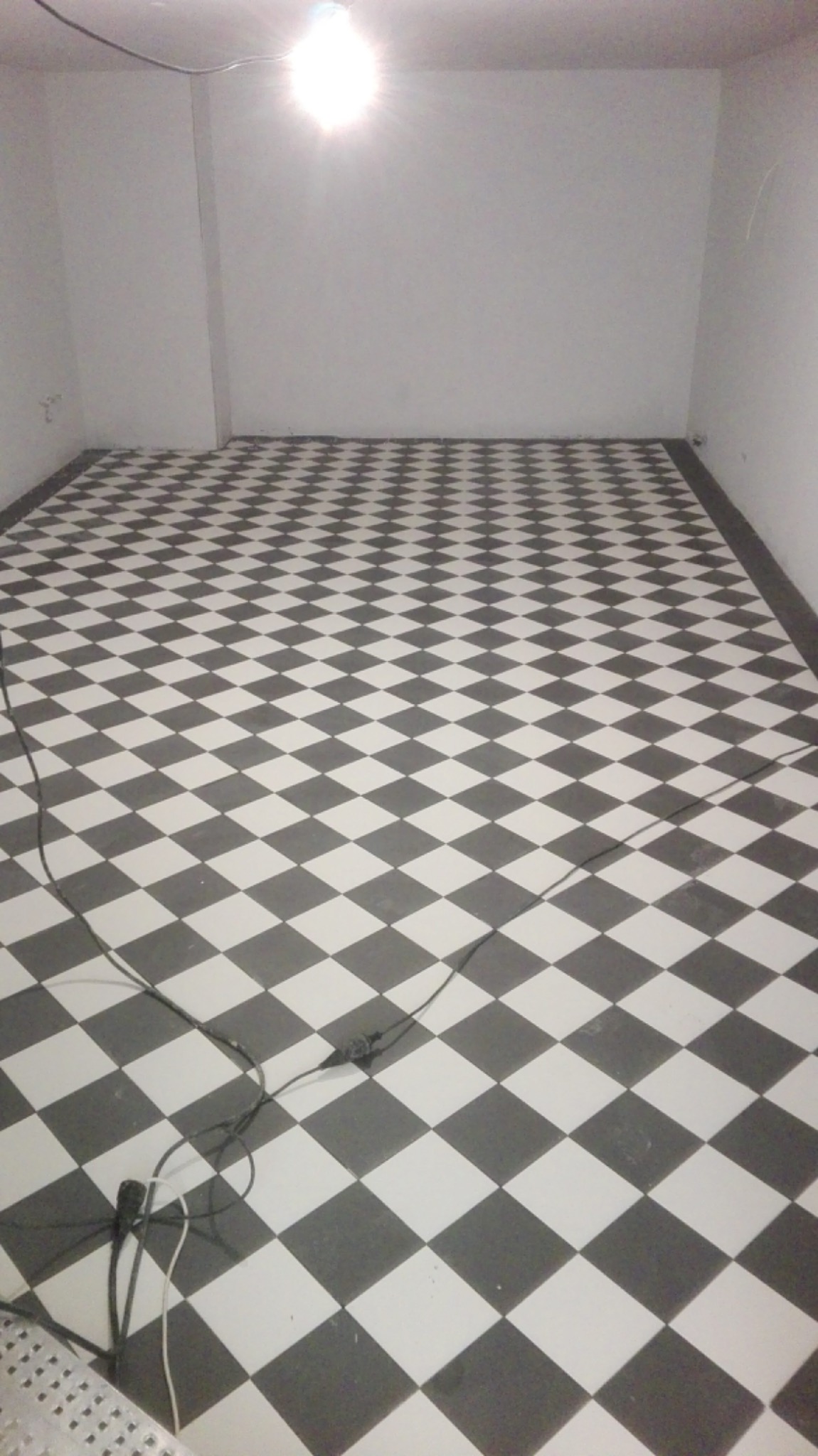 Nowo ułożona podłoga w szachownicę z czarno-białych płytek w pustym pomieszczeniu, widoczne kable elektryczne na podłodze.