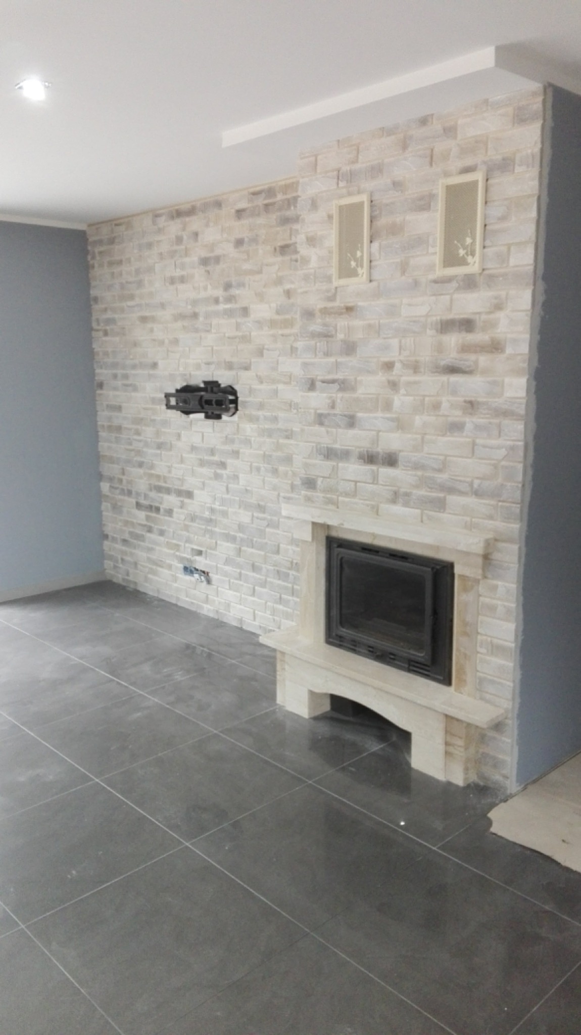 Ściana z cegły dekoracyjnej w odcieniach bieli i szarości z wbudowanym kominkiem, podwieszony uchwyt na telewizor, szara podłoga z płytek, dwa ozdobne panele ścienne