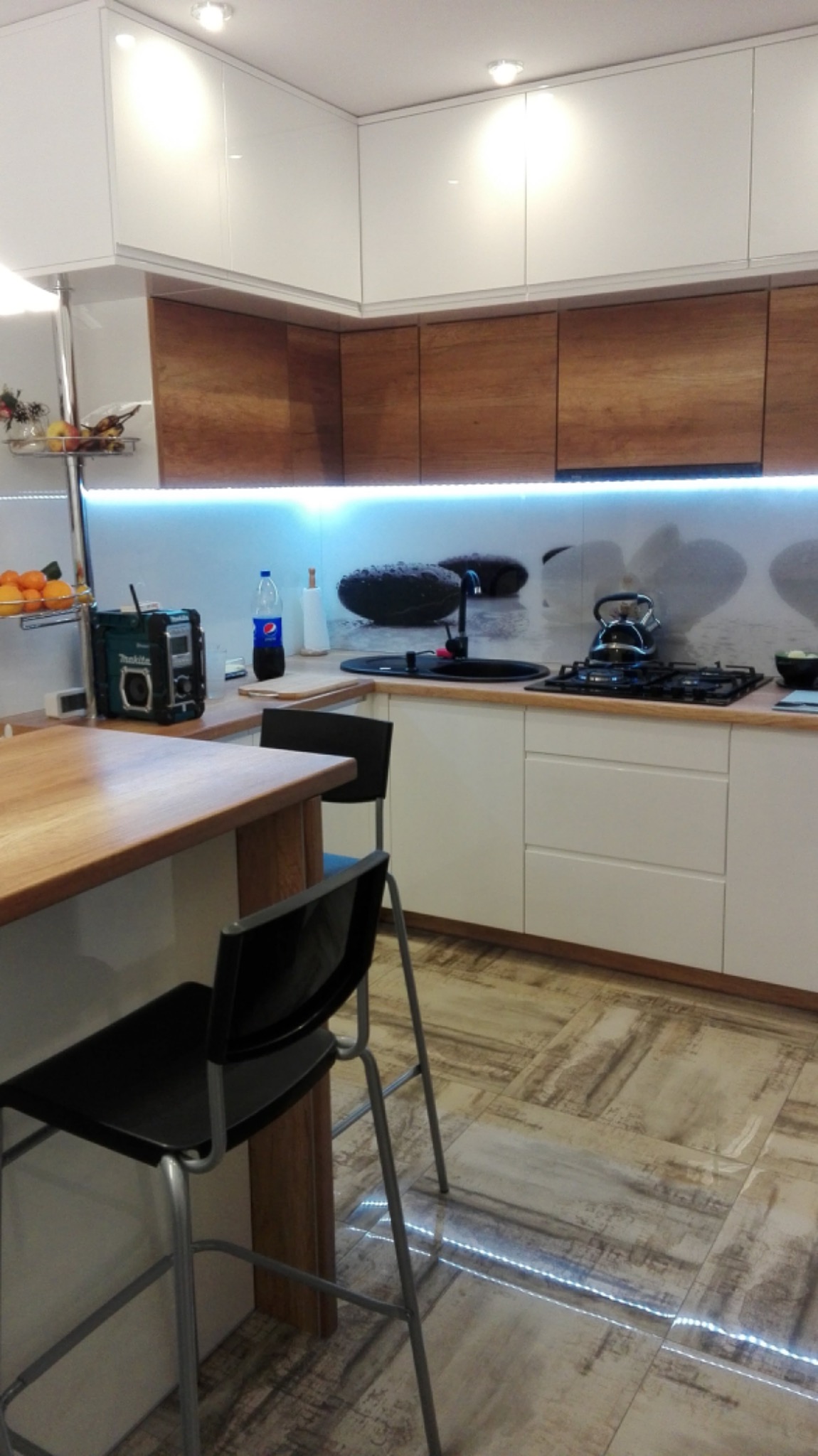 Nowoczesna kuchnia w Obornikach z białymi szafkami górnymi i drewnopodobnymi dolnymi, wyspą z drewnianym blatem i czarnymi hokerami. Na blacie kuchennym czajnik, płyta gazowa i czarny zlew, a...