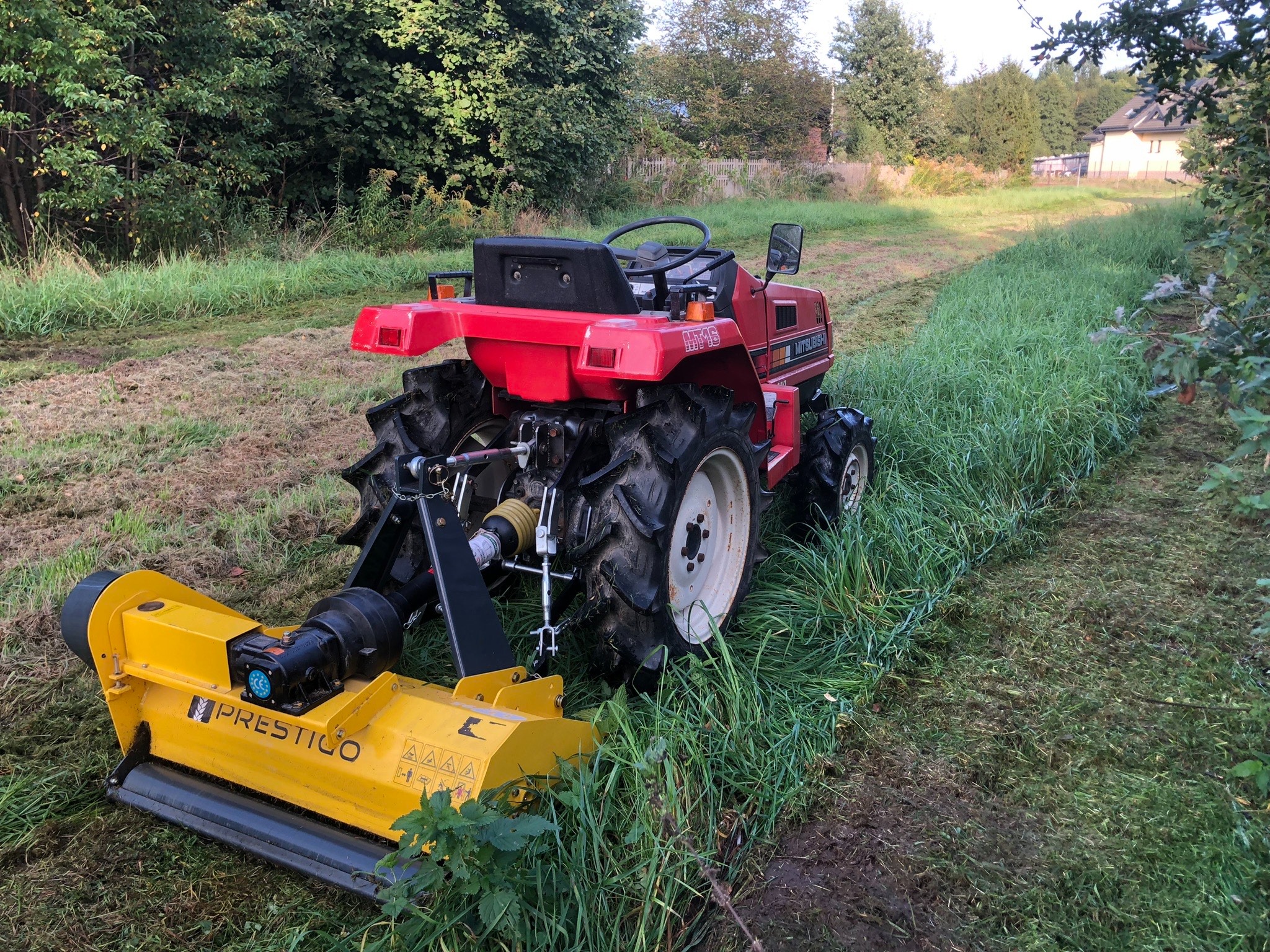 Czerwony traktor Mitsubishi z żółtą kosiarką bijakową Prestigio, widok z tyłu na częściowo skoszone pole z wysoką trawą, drzewa w tle.