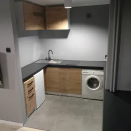 Mała kuchnia z drewnianymi frontami szafek, czarnym blatem, zlewem, pralką i małą lodówką, widziana z perspektywy narożnej.