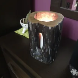 Drewniany świecznik wykonany z pnia drzewa z wkładem na świeczkę, oświetlony blaskiem płomienia, stojący na ciemnej komodzie obok ceramicznej miseczki z suszem.