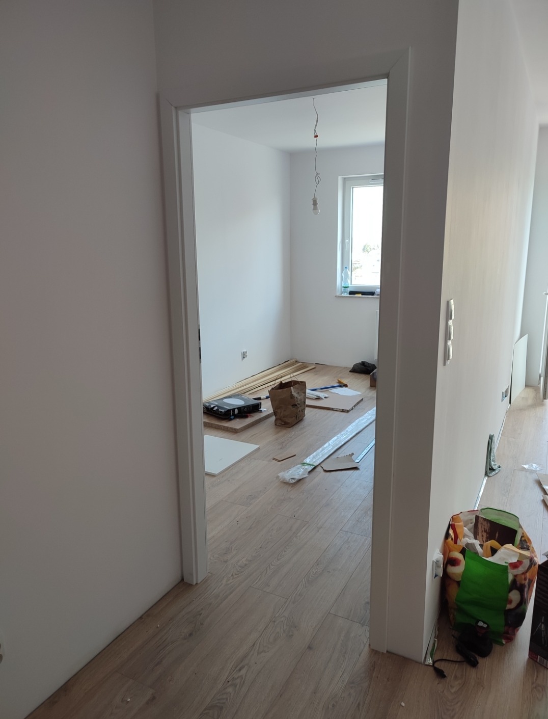Widok z korytarza na pokój w trakcie remontu, z widoczną podłogą wyłożoną panelami imitującymi drewno, narzędzia i materiały budowlane rozłożone na podłodze, oraz wiszącą żarówką bez klosza.