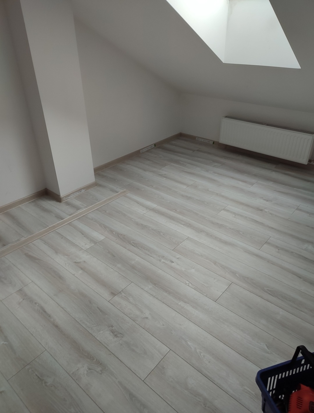 Jasne panele podłogowe ułożone w pokoju na poddaszu we Wrocławiu, widoczny grzejnik i okno dachowe, fragment koszyka w prawym dolnym rogu.