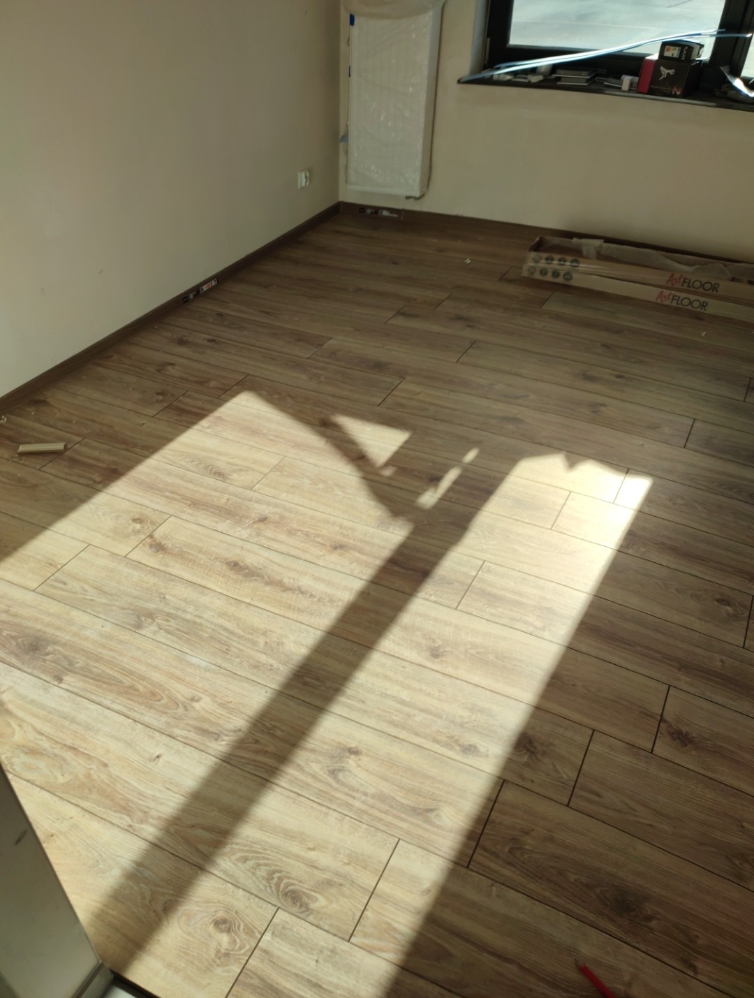 Jasne panele podłogowe ułożone w nowym pomieszczeniu, z widocznymi paczkami paneli przy oknie i długim cieniem padającym na podłogę.