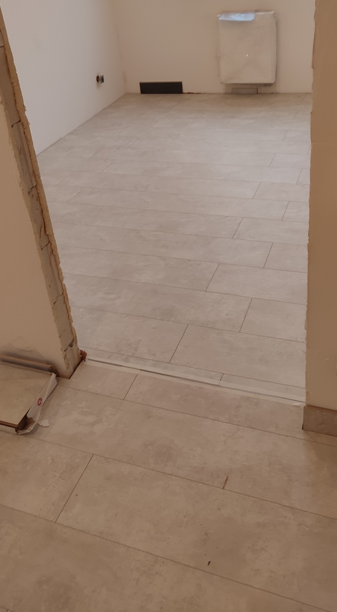 Ułożone panele podłogowe imitujące płytki ceramiczne w jasnym odcieniu, widoczny próg i fragment ściany w trakcie wykończenia.