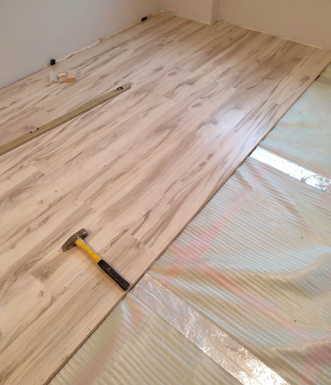 Fragment podłogi z jasnych paneli w trakcie montażu, widoczna poziomica, młotek z żółtą rękojeścią i folia izolacyjna z taśmą aluminiową.